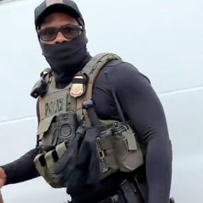 An agent wears a hat, sunglasses and black neck gaiter over his mouth, along with a body camera and a green vest that says “police.”