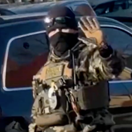 An agent wearing a camouflage uniform, black mask and black beanie waves to the camera. Only his eyes are visible.