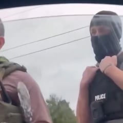 An agent wearing a black balaclava with a body camera and black vest with the word “police” is seen from inside a car window.